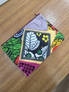 ACAP Made patchwork zipper pouch, front: black and white leaf wax-print panel with yellow flowers, lilac velvet corner, pink sequin net and green, red and orange wax prints, with a burgundy zip